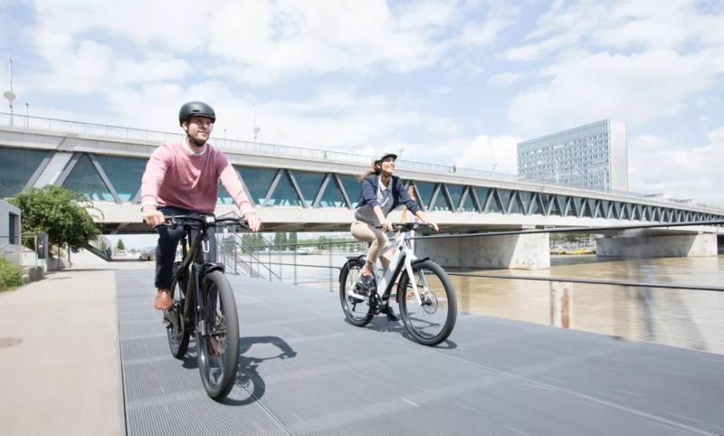 A woman and a man, both wearing bicycle helmets, are chatting and smiling. The woman is standing next to an e-bike, while the man is sitting on a bench with a racing bike next to him.